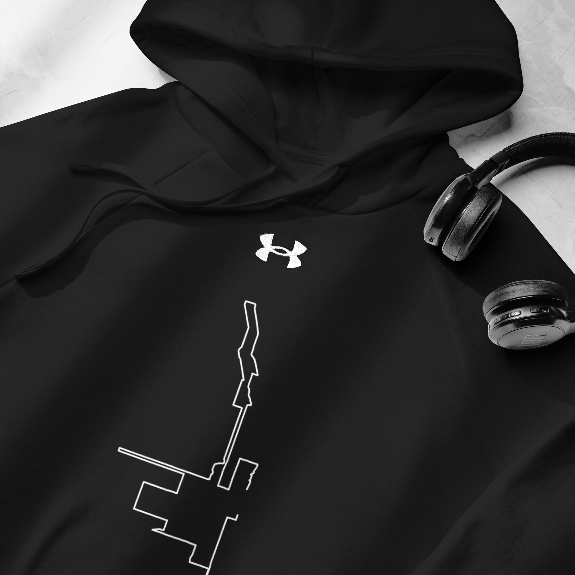 Black hoodie with Under Armour logo and white graphic design, paired with headphones on a light gray background