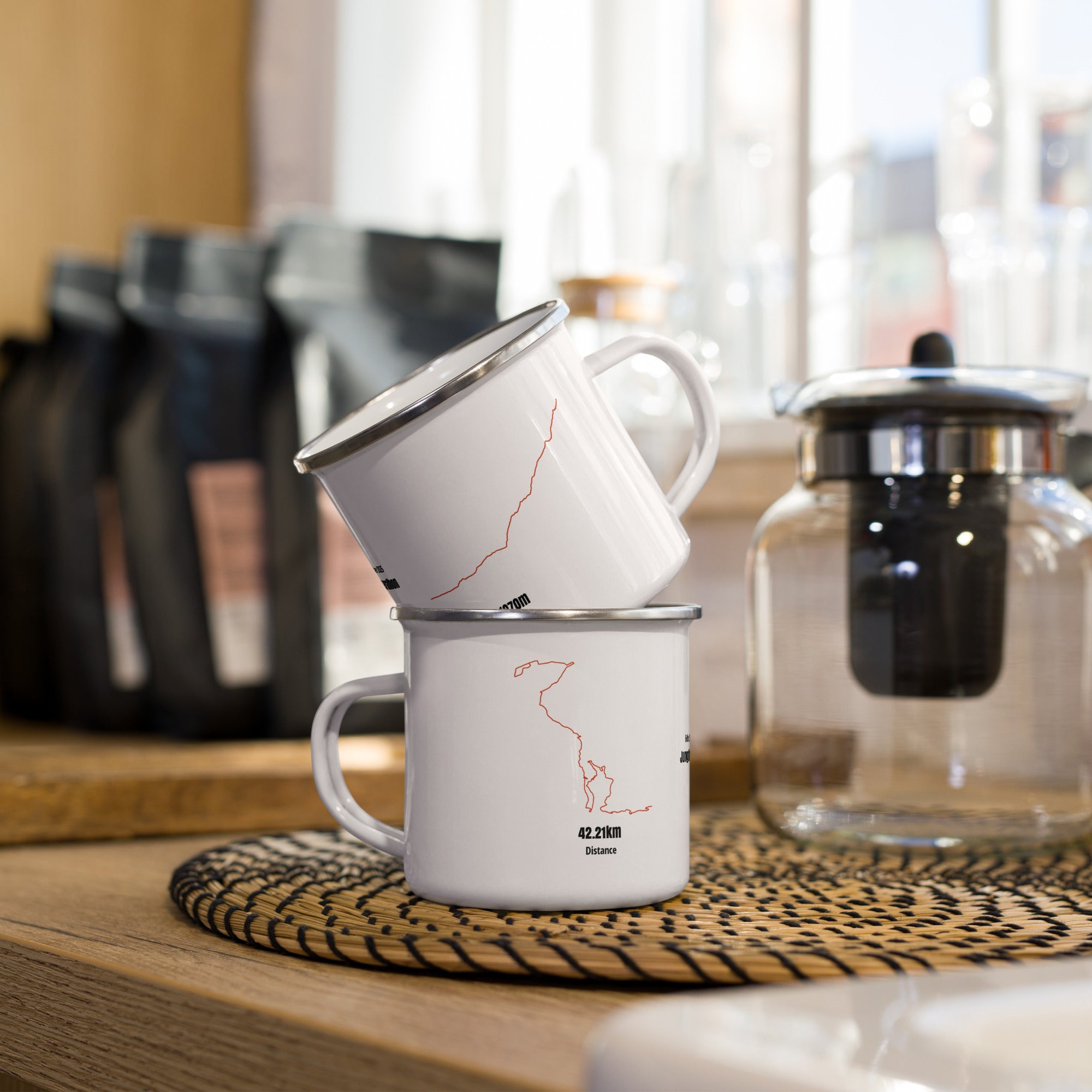 Two white mugs with red custom Strava route print designs stacked on a wooden surface with coffee-making equipment in the background.