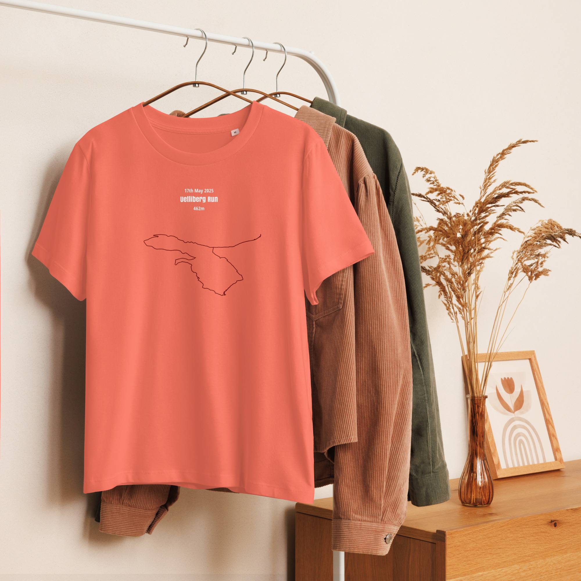 Coral t-shirt with a custom Strava route print design on a hanger against a neutral wall with a wooden surface