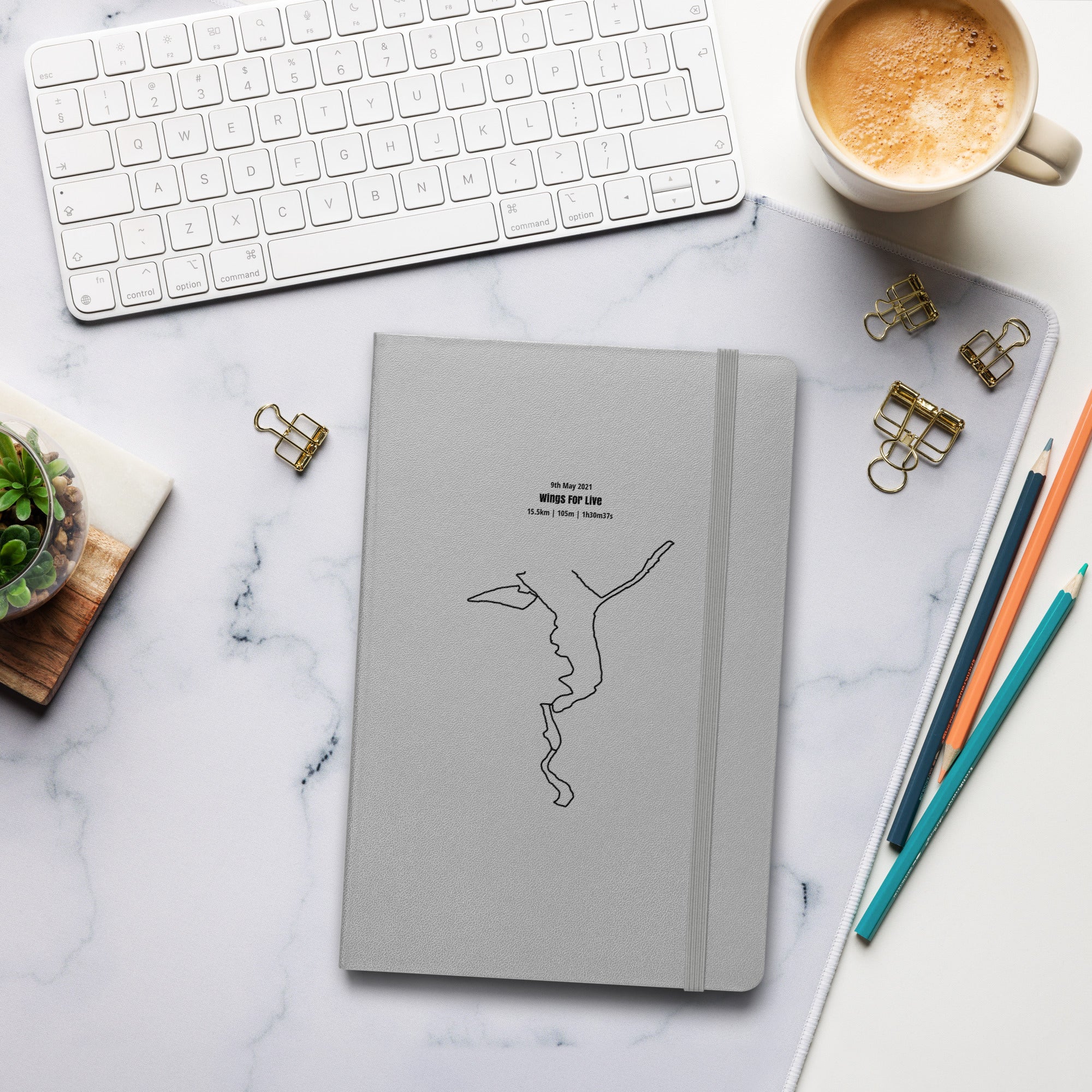 Gray notebook with custom Strava route print art on a marble desk with a keyboard and coffee cup