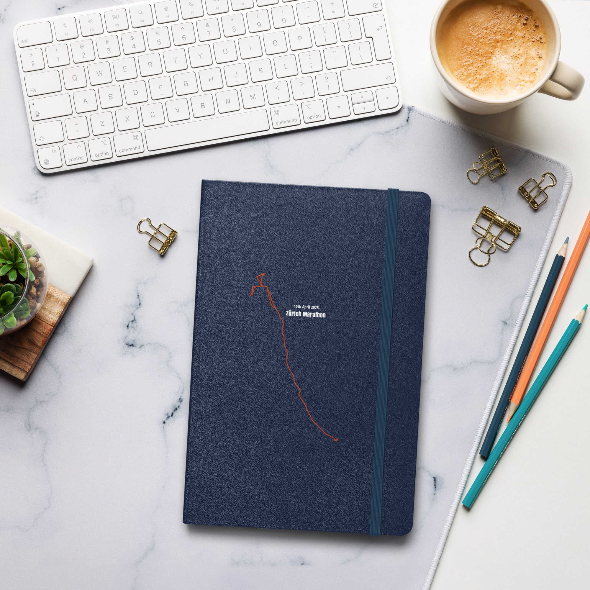 Blue notebook with custom Strava route print art on a marble desk with office supplies and a cup of coffee