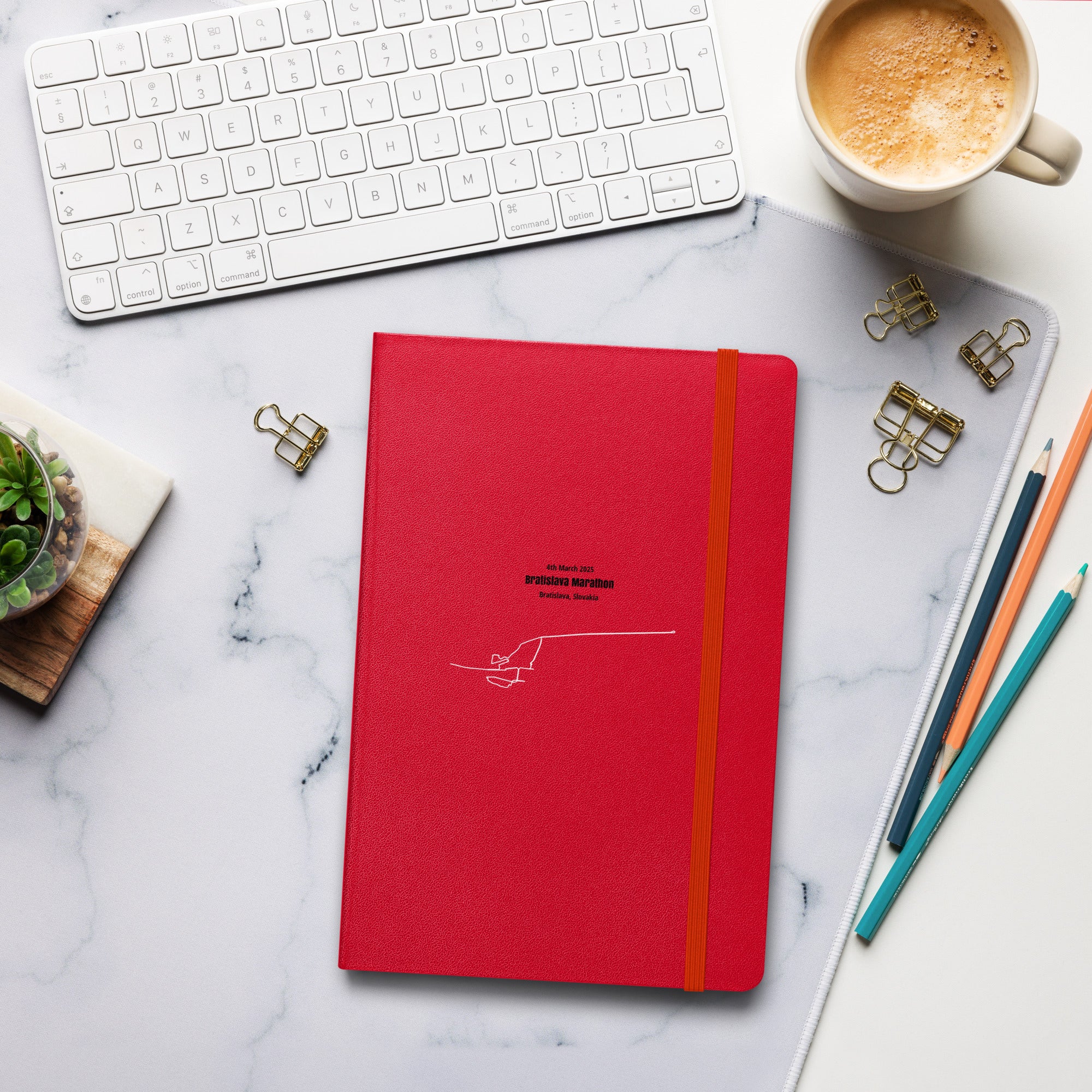 Red notebook with custom Strava route print art and an orange strap on a marble desk with office supplies and a cup of coffee