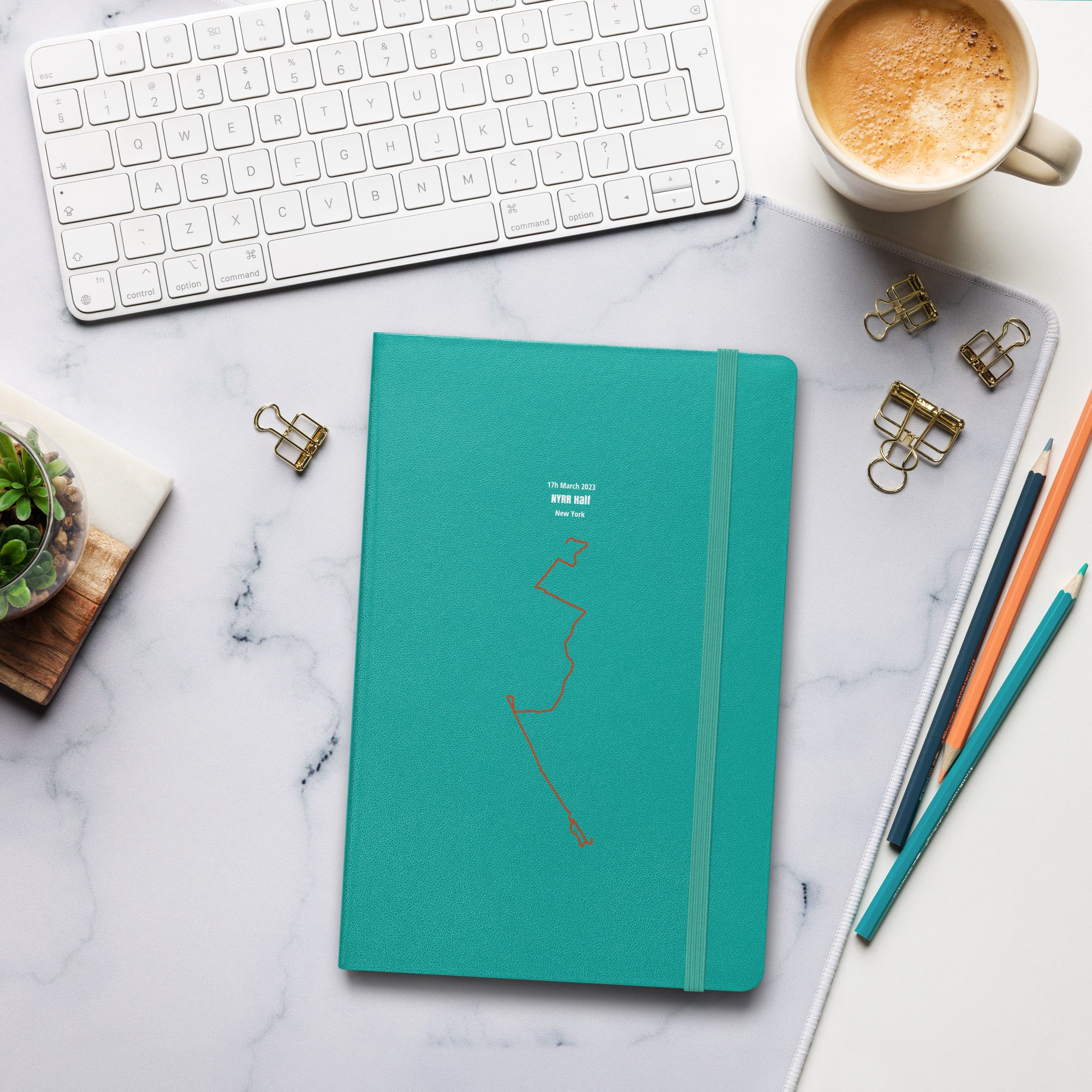 Teal notebook with custom Strava route print art on a marble desk with office supplies and a cup of coffee
