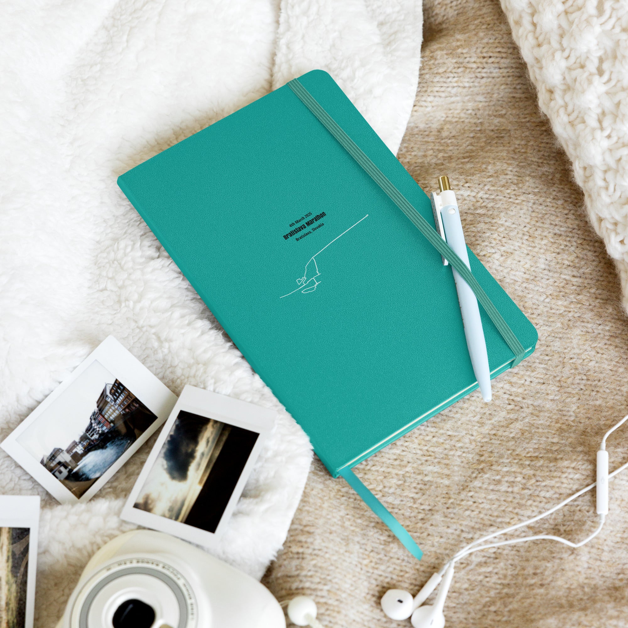 Teal notebook with custom Strava route print art and a pen on a textured surface with photos and earphones
